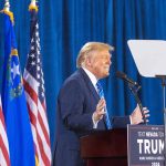 Person speaking at podium with flags in background