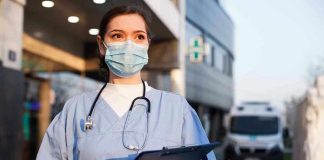 Nurse in scrubs and mask outside hospital holding clipboard