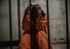 Person in orange jumpsuit sitting behind bars handcuffed