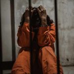 Person in orange jumpsuit sitting behind bars handcuffed