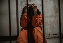 Person in orange jumpsuit sitting behind bars handcuffed
