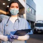 Nurse in scrubs and mask outside hospital holding clipboard.
