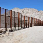 A long border wall stretches across a desert landscape with mountains in the background