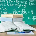 Open textbook on a desk with stacked books and a chalkboard filled with math equations in the background