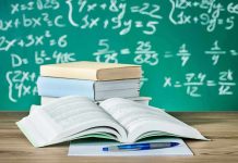 Open textbook on a desk with stacked books and a chalkboard filled with math equations in the background