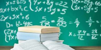 Open textbook on a desk with stacked books and a chalkboard filled with math equations in the background