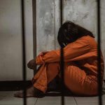 Reality Star FREED After Elderly Fraud Scheme Person in orange jumpsuit sitting behind bars, head down.