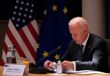 Biden’s Policies EXPOSED: Inflation Truth REVEALED Man seated at table with flags in background.