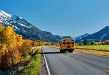 Electric School Buses Face Reality In Winter Weather A yellow school bus driving on a rural road surrounded by autumn trees and mountains