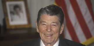 A smiling man in a suit with an American flag in the background