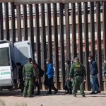 Border agents escort a group near a tall fence and a patrol van