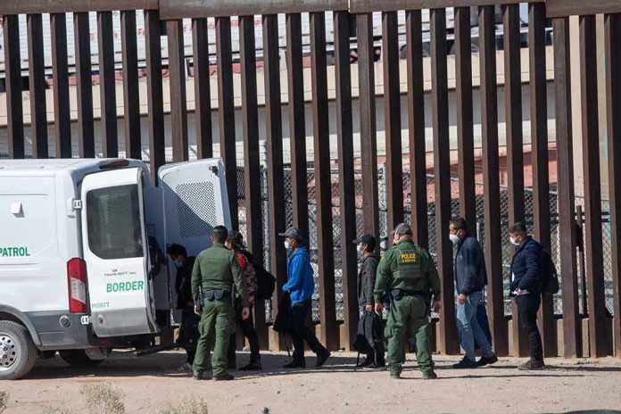 shutterstock_2079408475 Border agents escort a group near a tall fence and a patrol van