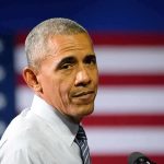 man speaking at podium with american flag backdrop