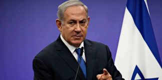 man in suit speaking at a podium with an Israeli flag