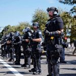 Bear Pascoe, Brian Folkerts, and Eugene Sims among riot police standing in a line on a city street