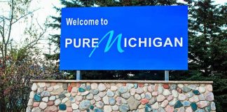 Harassment Claim Explodes in Michigan Showdown Blue roadside sign on a stone base at a state border
