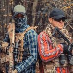 Two armed individuals in tactical gear standing back to back in a forest