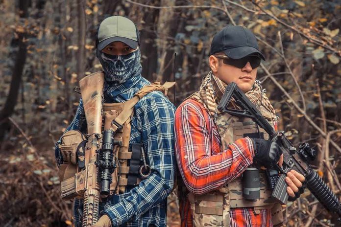 Two armed individuals in tactical gear standing back to back in a forest