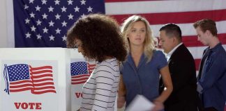 More Democrats Plan to Vote In-Person This Year Voters at polling booths in front of an American flag