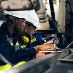 57 Illegals Taken Off the Line at a New York Factory Two factory workers in hard hats using a laptop near industrial machinery