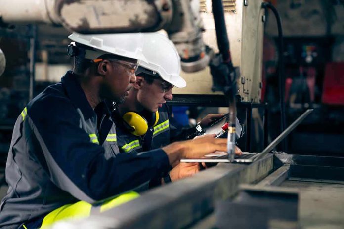 Two factory workers in hard hats using a laptop near industrial machinery