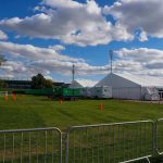 Temporary white tents and equipment on a grassy field with traffic cones and metal barriers