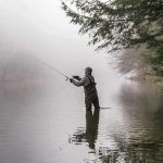Angler wading in a foggy river casting a fishing rod