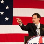 Speaker in suit pointing during speech before large American flag