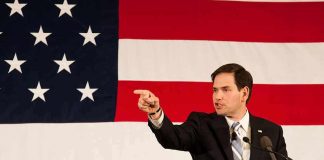 Rubio Defends National Loyalty Saying It Isn’t SELFISH Speaker in suit pointing during speech before large American flag